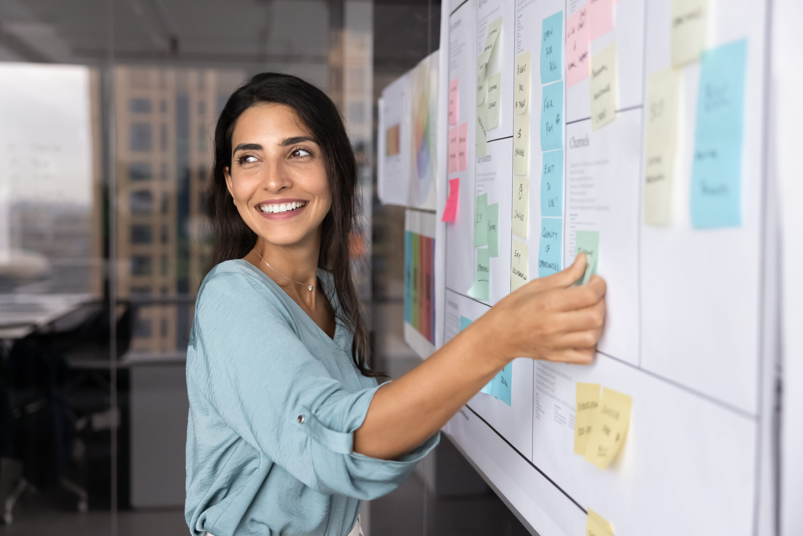 A woman smiling and placing sticky notes on a whiteboard covered with colorful notes and papers in a modern office setting.