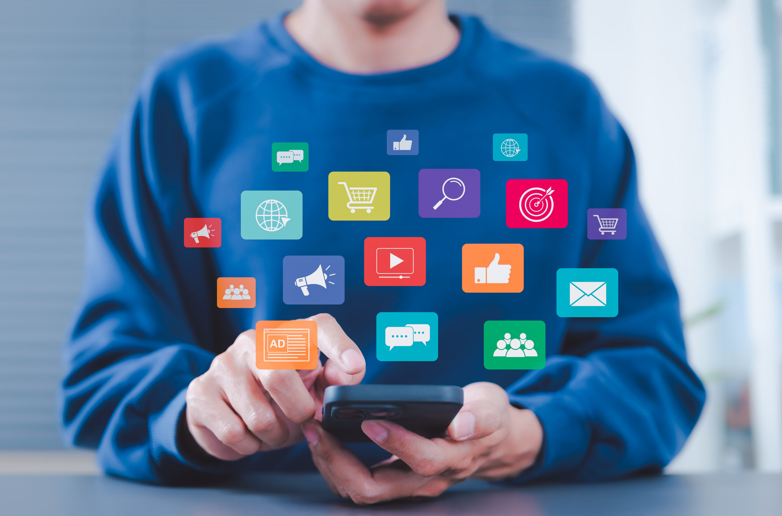 A person in a blue sweatshirt uses a smartphone. Colorful digital icons representing various online apps and social media functions, such as shopping, email, and messaging, appear above the phone.