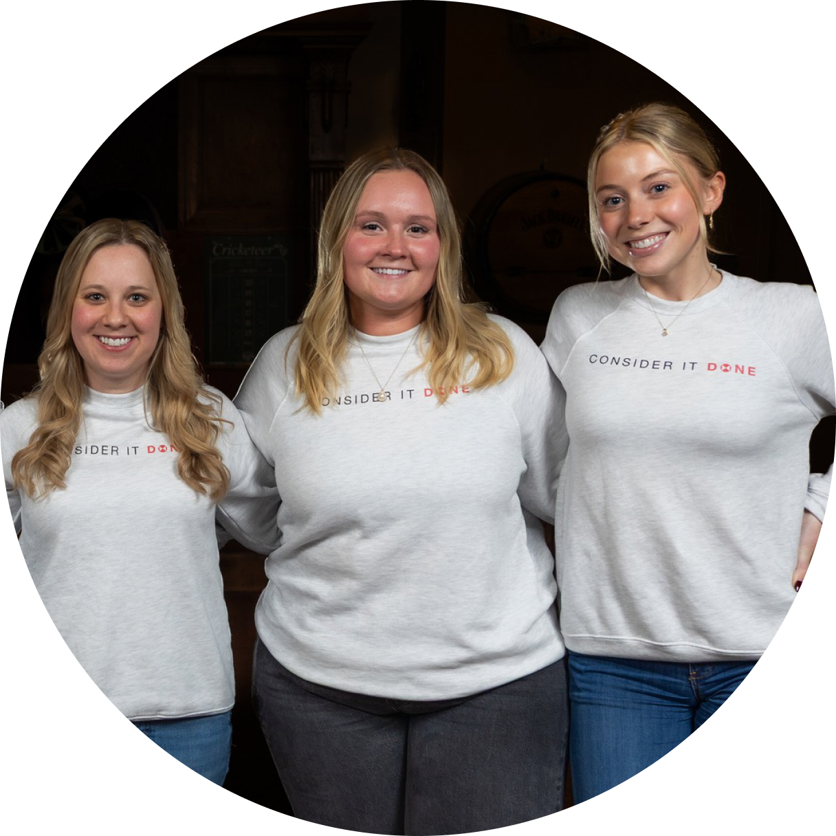 Three smiling women stand together, wearing matching light gray sweatshirts that say CONSIDER IT DONE in red lettering. They are posing indoors with arms around each other.